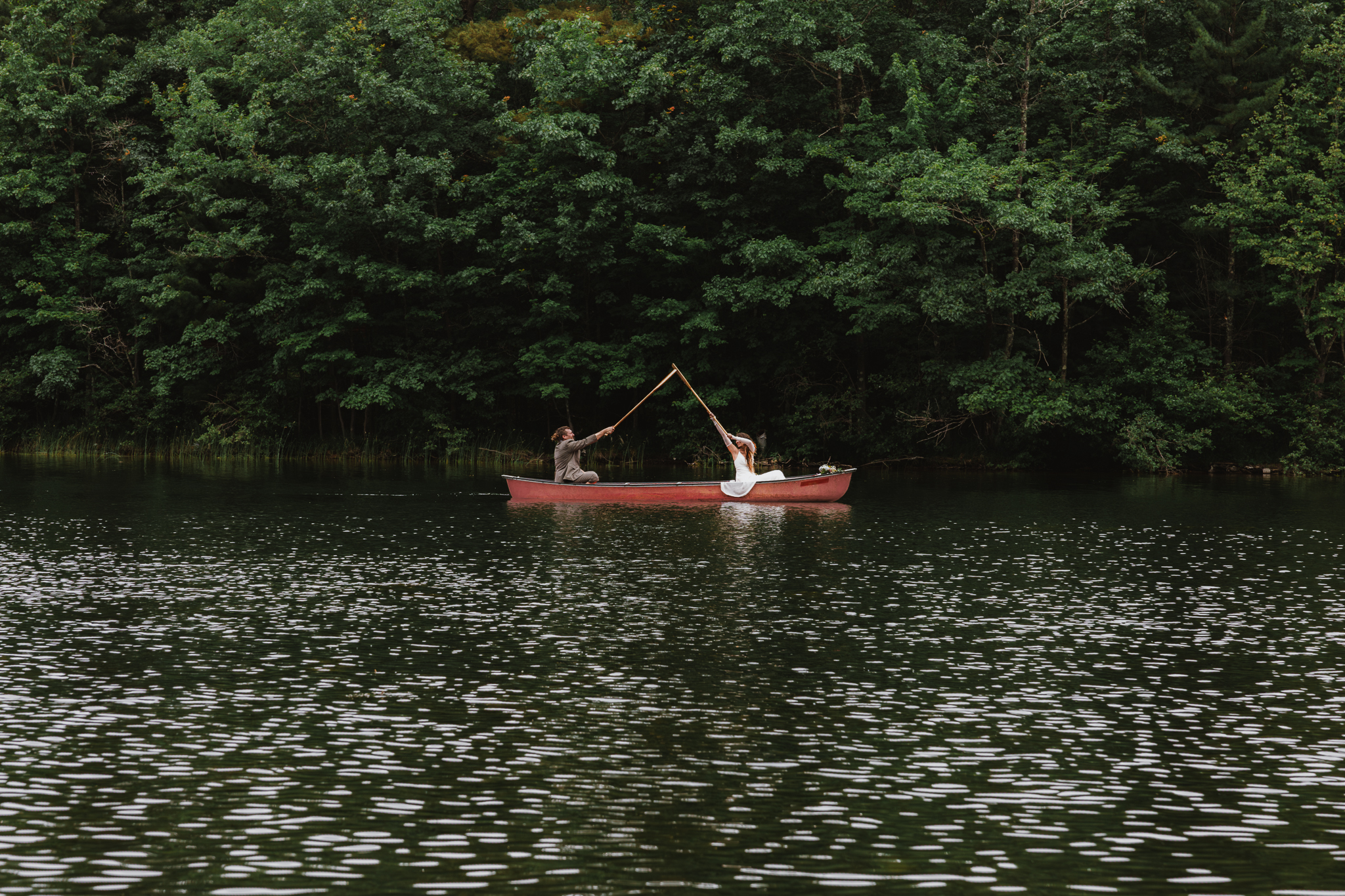 In the distance on the lake, a couple in red canoe holding their oars in their air celebrating their elopement. There is a thick tree line behind them.